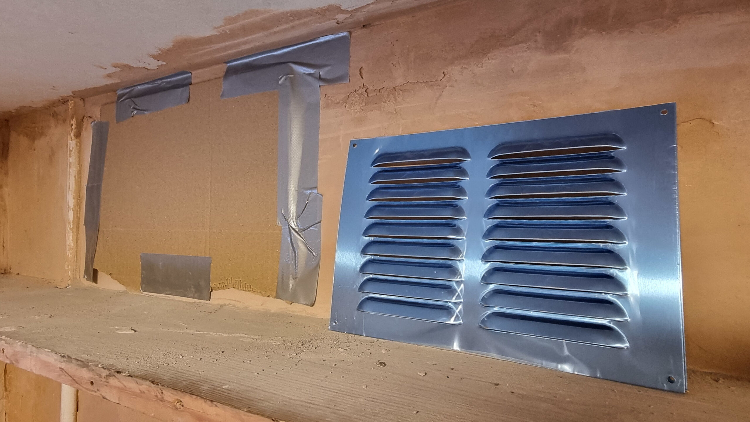 A cardboard cover is taped in place over the newly cut ventilation hole. The soon to be installed metal grille sits beside the hole on a shelf.